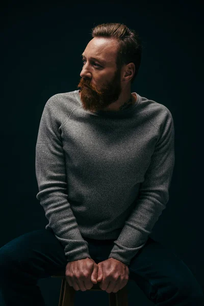 Fashionable man in sweater looking away while sitting on chair isolated on black with shadow