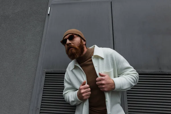 stylish man in beanie hat and sunglasses adjusting white shirt jacket outside