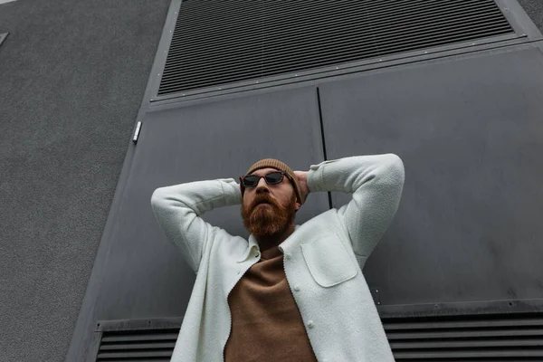 low angle view of stylish man in beanie hat and sunglasses standing in white shirt jacket and posing outside