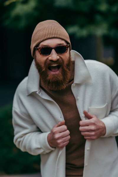 excited and bearded hipster in stylish sunglasses and beanie hat smiling outside 