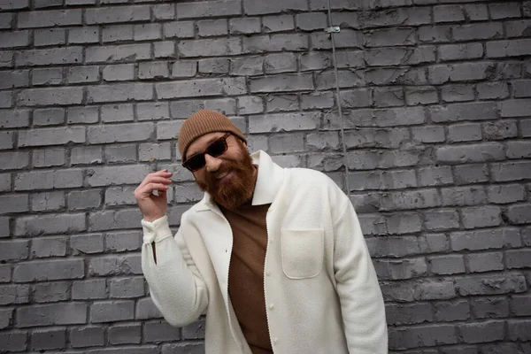 stock image happy and bearded man in beanie hat and sunglasses smiling near brick wall 