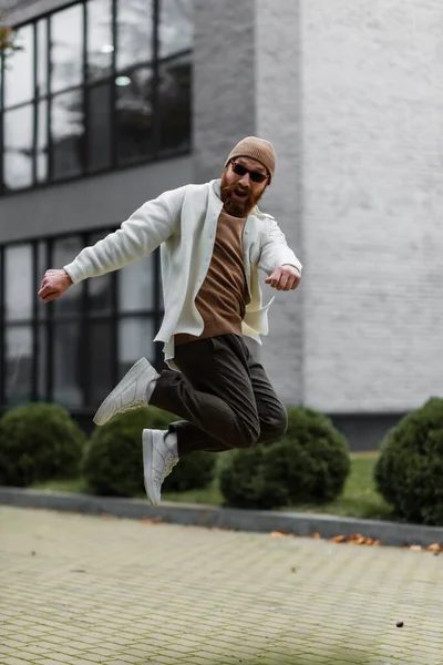 full length of excited man in stylish sunglasses and shirt jacket jumping near modern building