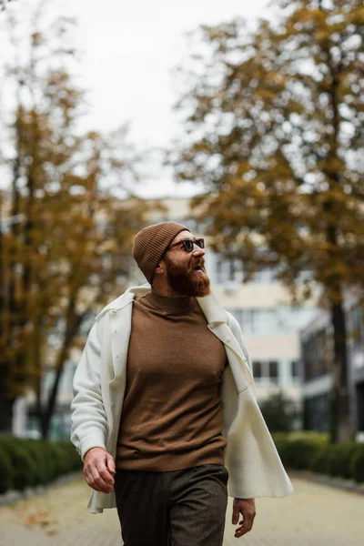 cheerful bearded man in beanie hat and trendy sunglasses looking away against trees and cloudy sky