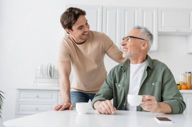 Mutlu genç adam gülümseyen babaya bakıyor. Elinde kahve fincanı, yanında boş ekranlı akıllı telefon oturuyor.