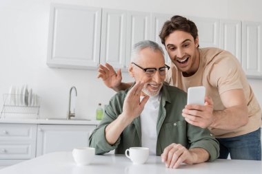 Mutfaktaki cep telefonuyla video görüşmesinde yetişkin oğlu olan neşeli bir adam el sallıyor.