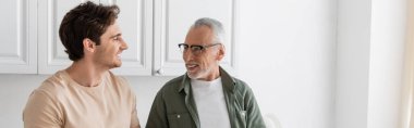 positive man looking at grey haired dad in eyeglasses during conversation in kitchen, banner clipart