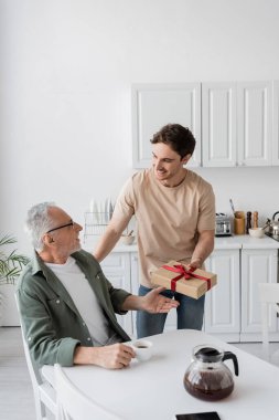 smiling man presenting fathers day gift to pleased dad sitting in kitchen with coffee cup clipart