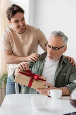 amazed man in eyeglasses holding fathers day present near smiling son clipart