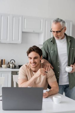 Elinde kahve fincanı olan olgun bir adam mutfakta dizüstü bilgisayarın yanında gülümseyen küçük oğlunun omzuna dokunuyor.