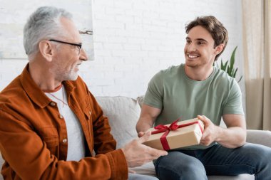 happy man congratulating dad on fathers day and giving him gift box on couch at home clipart