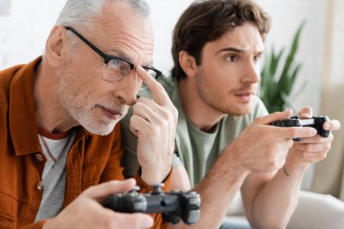 KYIV, UKRAINE - MAY 11, 2022: grey haired man adjusting eyeglasses while playing video game with young son  clipart