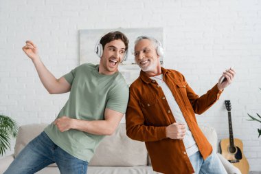 Kablosuz kulaklıklı heyecanlı adam evde olgun ve neşeli babasıyla şarkı söyleyip dans ediyor.