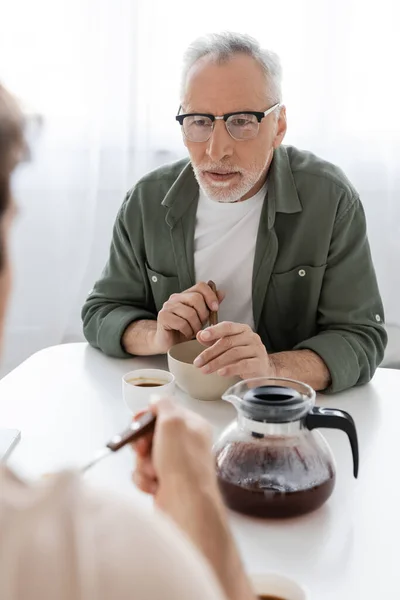 attentive man in eyeglasses looking at blurred son near breakfast and coffee pot on kitchen table