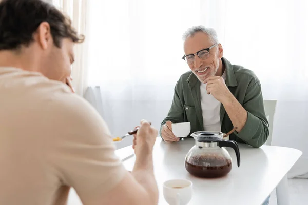 happy man in eyeglasses looking at blurred son having breakfast near coffee pot on kitchen table