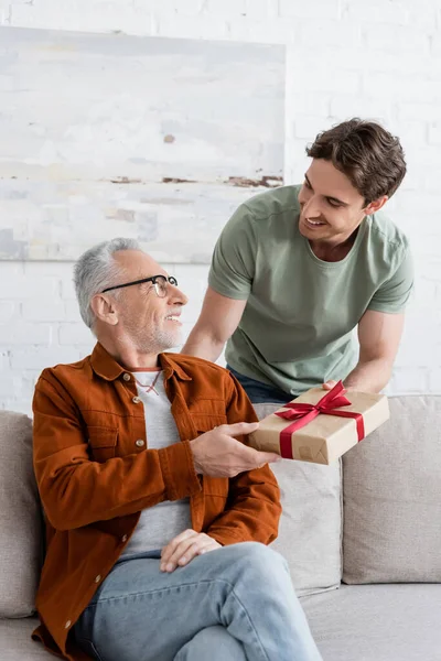 cheerful young man presenting fathers day gift to dad sitting on sofa in living room
