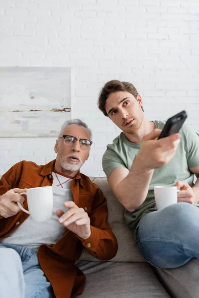 tensed dad and son with tea cups watching tv on sofa in living room