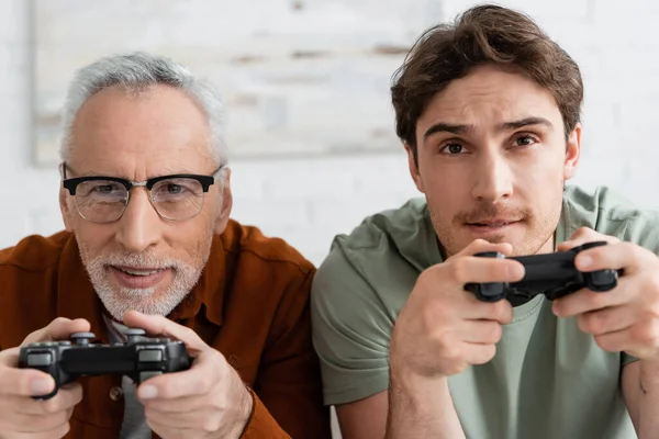 stock image KYIV, UKRAINE - MAY 11, 2022: tensed guy with joystick playing video game with smiling grey haired dad