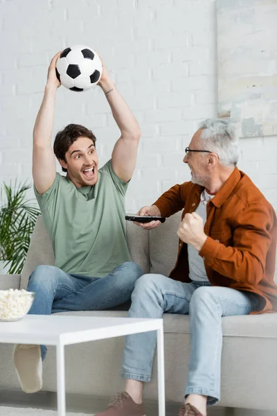 Stock image cheerful man shouting and holding soccer ball in raised hands near mature dad showing win gesture 