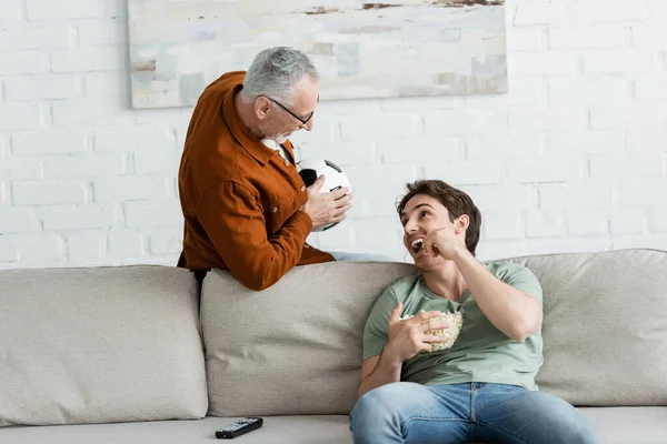happy man eating popcorn on couch and looking at dad holding soccer ball in living room