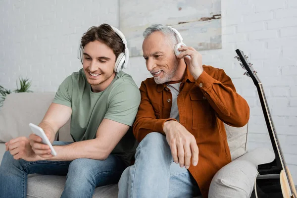 Stock image smiling father and son in wireless headphones looking at cellphone while sitting in living room