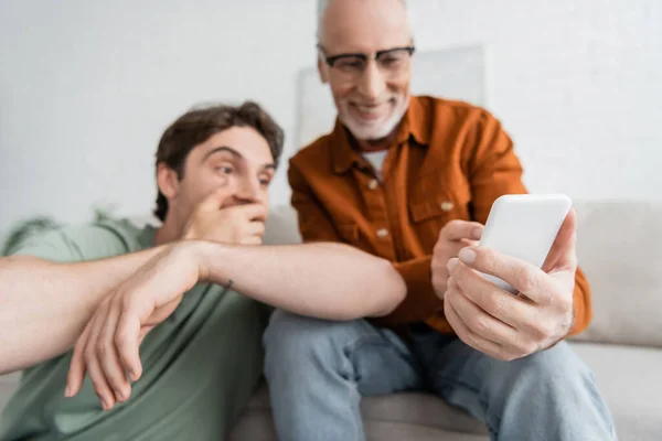 happy and mature father with beard holding smartphone near young son on blurred background