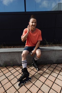 Smiling man in earphone and roller blades holding coffee to go on urban street  clipart