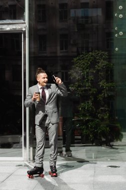 amazed businessman in roller skates looking away while standing with smartphone and paper cup near glass building clipart