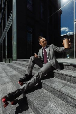 Takım elbiseli ve patenli gülümseyen işadamı merdivenlerde oturmuş akıllı telefon ve kahveyle dışarıyı seyrediyor.