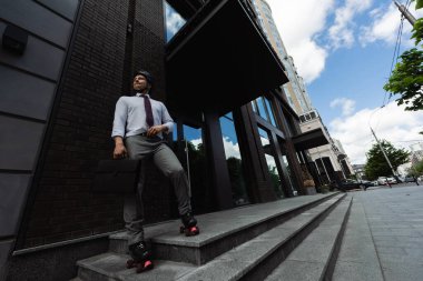 full length of man in formal wear and roller skates holding briefcase and looking away on urban street clipart