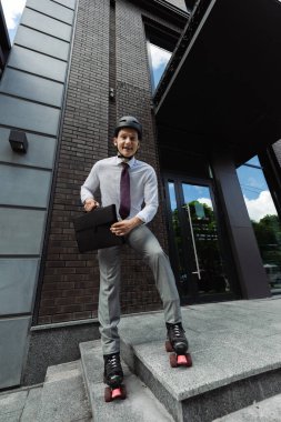 cheerful roller skater in formal wear and helmet holding briefcase and smiling at camera near modern building clipart