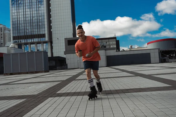 Cheerful man in roller skates holding smartphone and coffee to go on city street