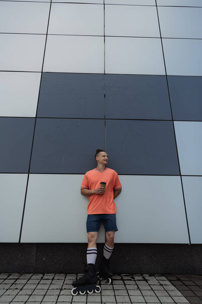 Wide angle view of smiling roller skater holding coffee to go near building outdoors 