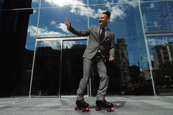 happy businessman in grey suit waving hand while rollerblading on urban street near modern building