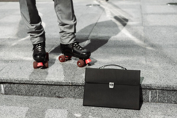 black briefcase near cropped businessman in roller skates on sidewalk of city street