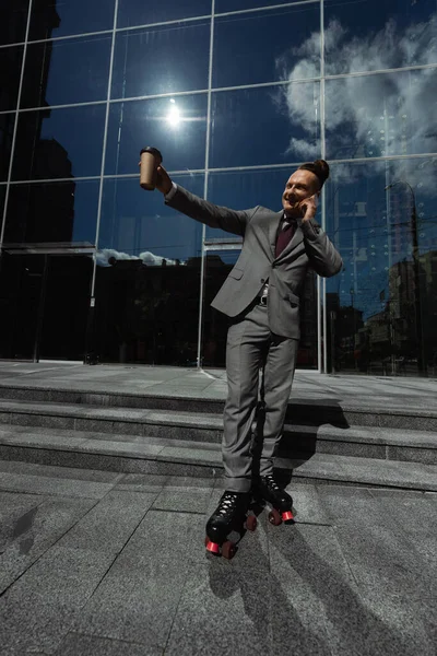 スーツ姿の幸せなビジネスマンとスマートフォンで話してるローラースケートストリートで紙コップを手に持ってる — ストック写真