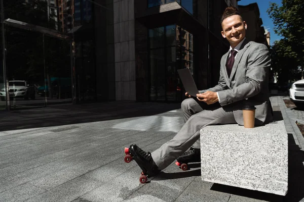 smiling man in roller skates and formal wear looking at camera while sitting with laptop on street