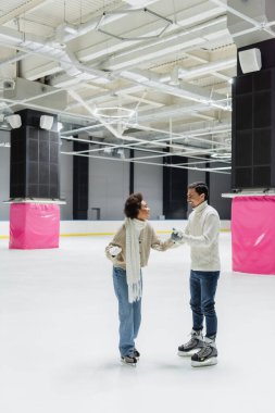 Gülümseyen Afro-Amerikalı kadın, erkek arkadaşının elini tutarken buz pateni yaparken görülüyor. 