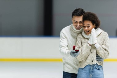 Young man in sweater holding engagement ring and hugging african american girlfriend on ice rink  clipart