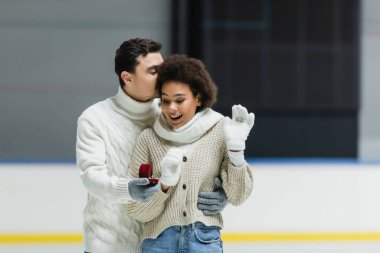Man in sweater kissing african american girlfriend and holding proposal ring on ice rink  clipart