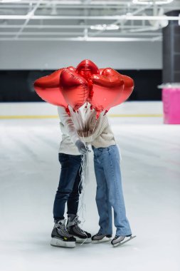 Young couple in warm clothes holding balloons in shape of heart on ice rink  clipart