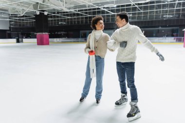 Smiling african american woman holding gift box and looking at boyfriend on ice rink  clipart