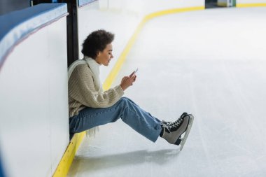 Buz pateni pistinde akıllı telefon kullanan Afro-Amerikan kadının yan görüntüsü 