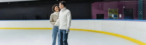 穿着毛衣微笑着的非洲裔美国女人看着男朋友在冰球场上 一面横幅 — 图库照片
