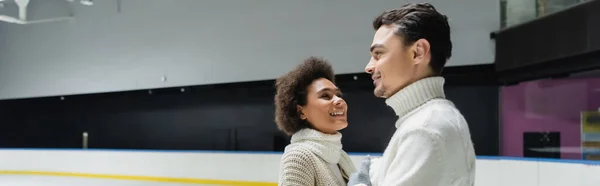 穿着围巾的积极的非洲女人看着男朋友在冰球场上 横幅上 — 图库照片