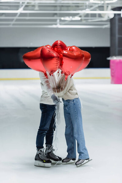 Young couple in warm clothes holding balloons in shape of heart on ice rink 