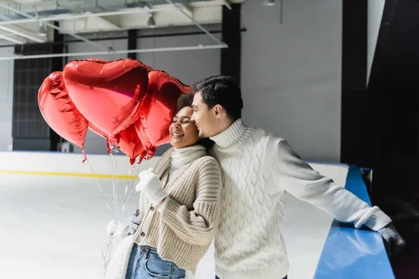 Pleased man in sweater hugging african american girlfriend with heart shaped balloons on ice rink
