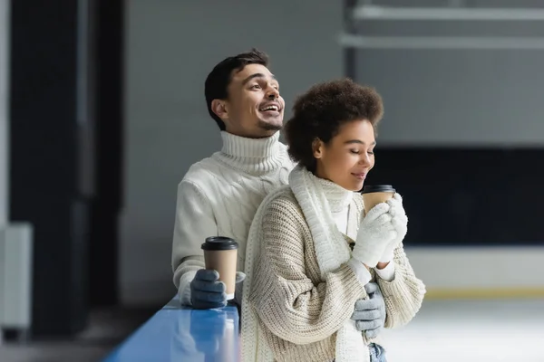 Smiling man in sweater holding coffee to go and hugging african american girlfriend on ice rink