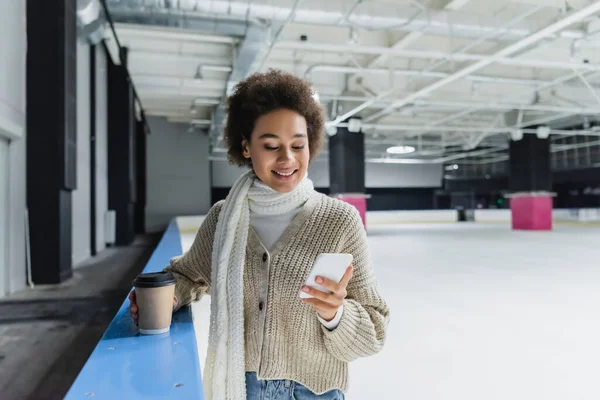 Smiling african american woman in scarf using smartphone and holding coffee to go on ice rink