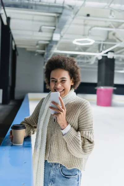拿着智能手机和纸杯在冰球场上微笑的非洲裔美国女人 — 图库照片