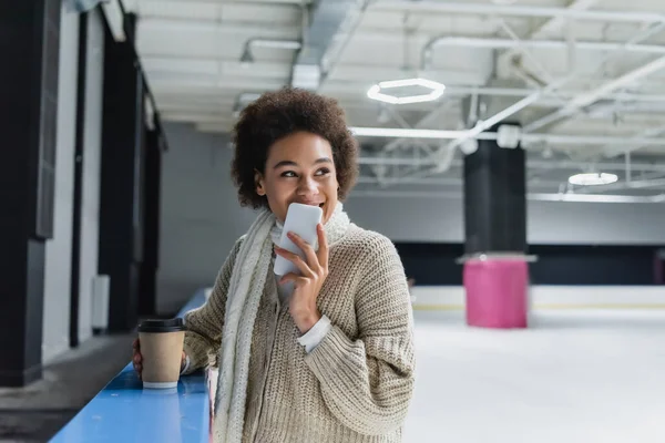 Young african american woman in warm scarf holding smartphone and coffee to go on ice rink